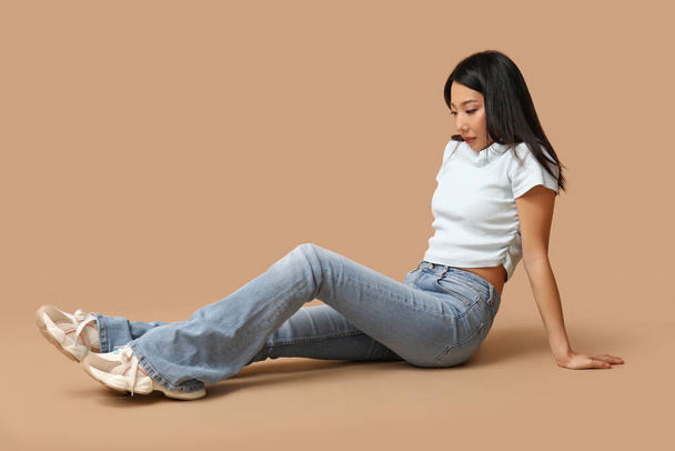 Красивая молодая азиатка в стильных джинсах сидит на коричневом фоне - Фото, изображение