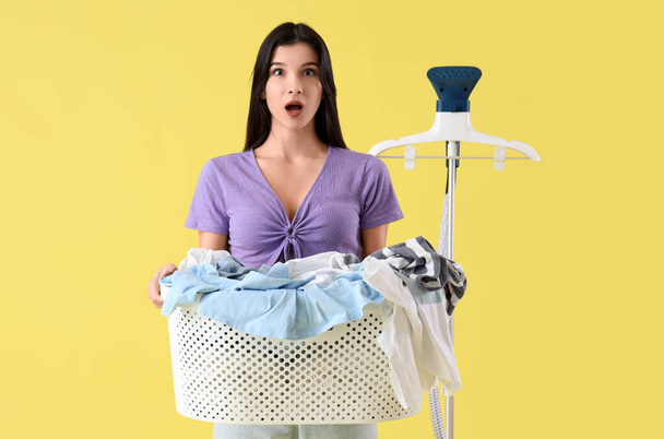 Shocked young woman with modern garment steamer and basket full of laundry on yellow background - Photo, Image