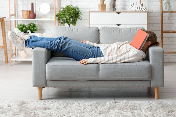 Όμορφη νεαρή γυναίκα με βιβλίο που βρίσκεται σε άνετο καναπέ στο σαλόνι στο σπίτι - Φωτογραφία, εικόνα
