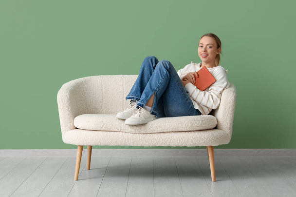Молодая счастливая женщина читает книгу на удобном диване у зеленой стены - Фото, изображение