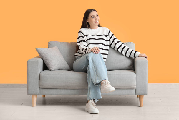 Красива молода щаслива жінка сидить на зручному дивані біля жовтої стіни - Фото, зображення