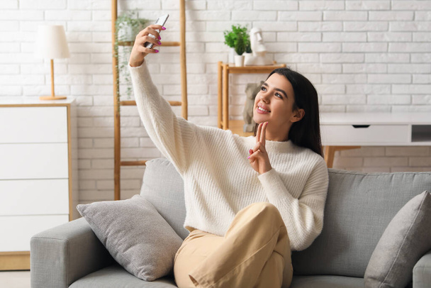 Güzel, genç ve mutlu bir kadın rahat bir koltukta oturuyor ve evdeki oturma odasında selfie çekiyor. - Fotoğraf, Görsel