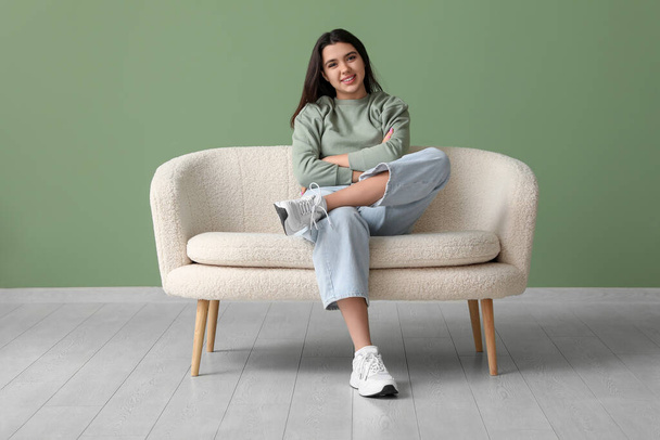 Όμορφη νεαρή ευτυχισμένη γυναίκα κάθεται σε άνετο καναπέ κοντά στο πράσινο τοίχο - Φωτογραφία, εικόνα