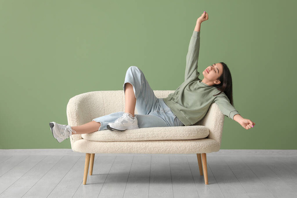 Όμορφη νεαρή γυναίκα χαλαρώνοντας σε άνετο καναπέ κοντά στο πράσινο τοίχο - Φωτογραφία, εικόνα