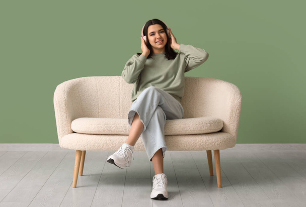 Gyönyörű fiatal boldog nő fülhallgató ül kényelmes kanapén, és zenét hallgatni közel zöld fal - Fotó, kép