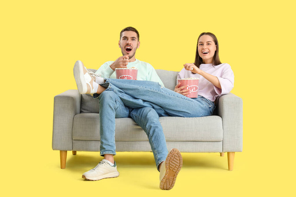 Mooi jong gelukkig paar met emmers popcorn kijken TV op de bank tegen gele achtergrond - Foto, afbeelding