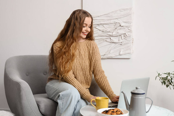 Mujer joven con taza de té en sillón utilizando el ordenador portátil en casa - Foto, imagen