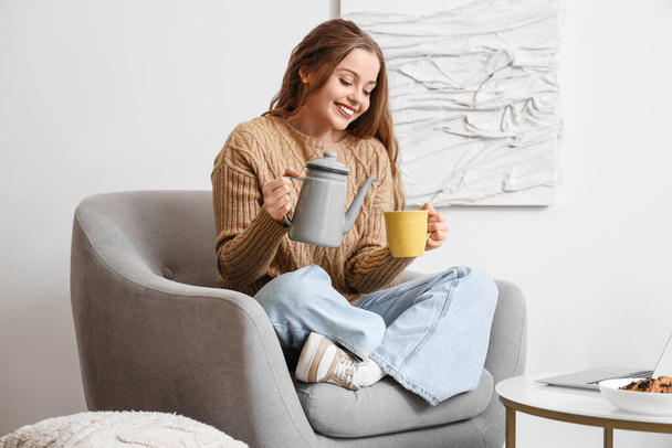Young woman pouring tea into cup in armchair at home - Photo, Image