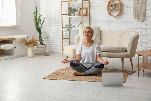 Reife Frau mit Laptop meditiert zu Hause. Online Yoga-Kurse - Foto, Bild