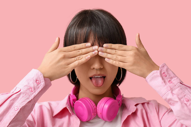 Mujer joven con estilo con piercing en la nariz y auriculares que muestran la lengua en el fondo rosa - Foto, imagen