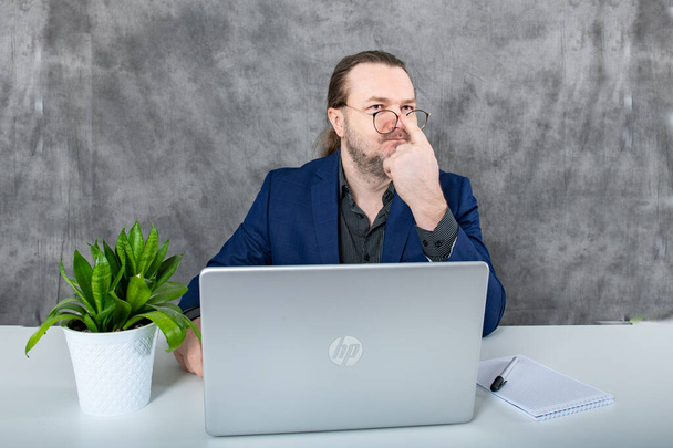 Een zakenman in een elegant blauw pak zittend aan een bureau in een eigentijdse kantooromgeving, met een laptop met een groene plant op de achtergrond, die een vleugje natuur toevoegt aan de werkruimte. - Foto, afbeelding