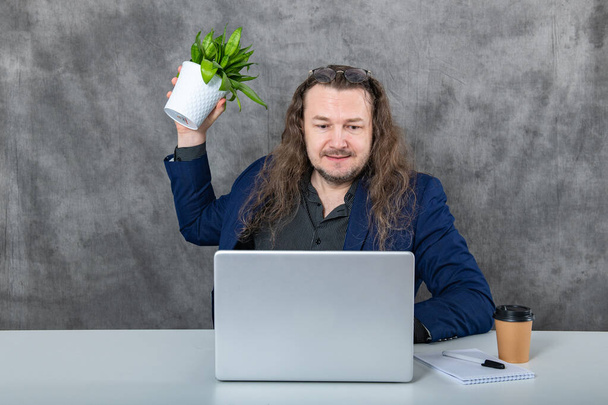 Um jovem e ambicioso empresário com cabelos longos sentado em um laptop e segurando uma planta verde enquanto trabalhava em um ambiente de escritório contemporâneo. - Foto, Imagem