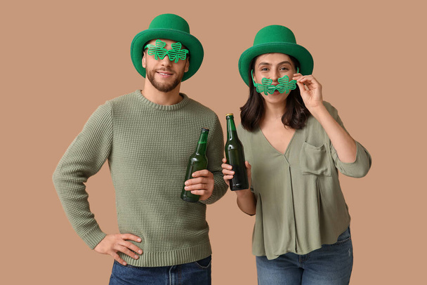 Beau jeune couple en lutin chapeaux et verres décoratifs en forme de trèfle avec des bouteilles de bière sur fond brun. Célébration de la Saint Patrick - Photo, image