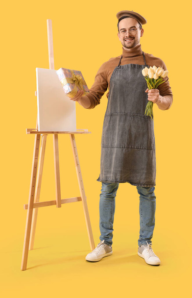 Ευτυχισμένος αρσενικό καλλιτέχνη με καβαλέτο, κουτί δώρου και μπουκέτο τουλίπες σε κίτρινο φόντο. Διεθνής Ημέρα της Γυναίκας - Φωτογραφία, εικόνα
