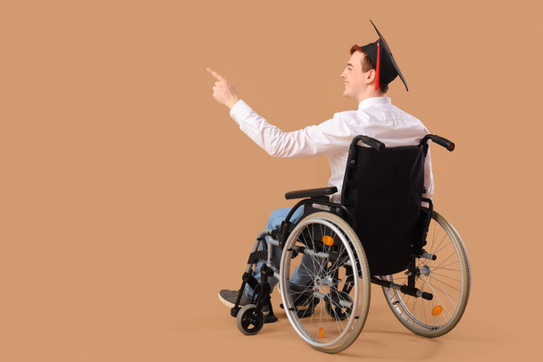 Masculino graduado em cadeira de rodas apontando para algo em fundo bege - Foto, Imagem