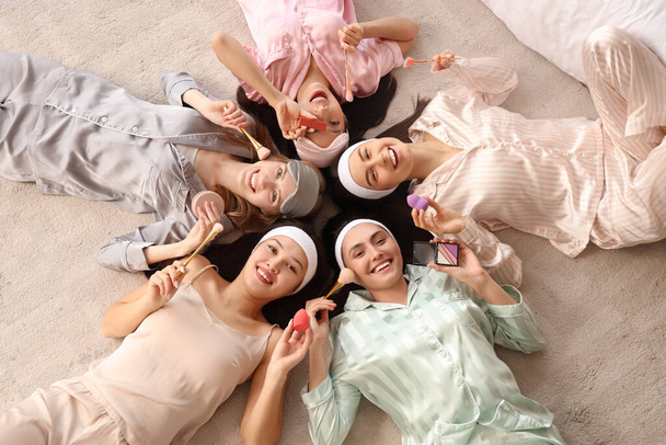 Mujeres jóvenes con productos de maquillaje tumbados en el suelo en la despedida de soltera en el dormitorio, vista superior - Foto, imagen