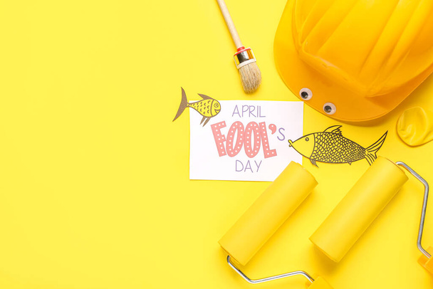 Hoja con texto ABRIL FOOL 'S DAY, herramientas de construcción y peces de papel sobre fondo amarillo - Foto, Imagen