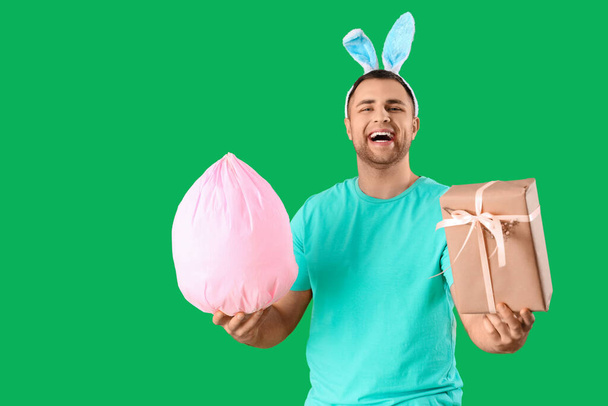 Щасливий молодий чоловік у вухах кролика з подарунковою коробкою та яйцем на зеленому фоні. Великоднє святкування - Фото, зображення