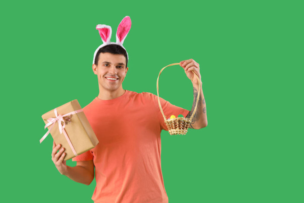 Joven feliz en orejas de conejo con caja de regalo y canasta de mimbre sobre fondo verde. Celebración Pascua - Foto, Imagen