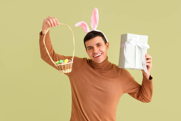 Szczęśliwy młody człowiek w króliczych uszach z pudełkiem prezentów i wiklinowym koszem na zielonym tle. Wielkanoc - Zdjęcie, obraz
