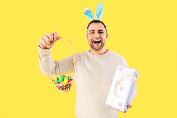 Joven feliz en orejas de conejo con caja de regalo y canasta de mimbre sobre fondo amarillo. Celebración Pascua - Foto, Imagen