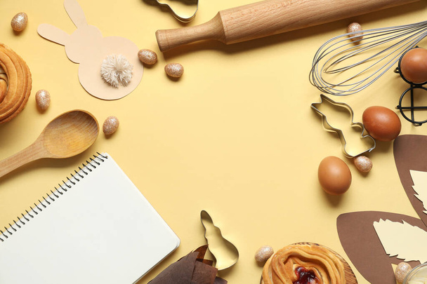 Marco de bloc de notas en blanco para recetas con bollos dulces, decoración de Pascua y herramientas para hornear sobre fondo amarillo - Foto, Imagen