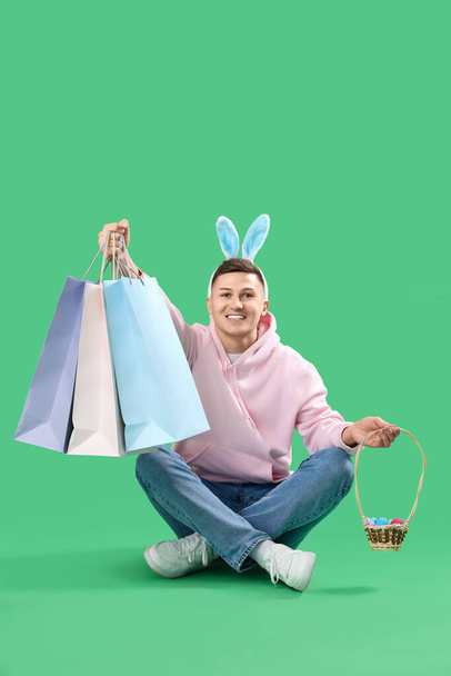 Fiatal férfi nyuszifül bevásárló táskák és húsvéti tojás ül zöld háttér - Fotó, kép