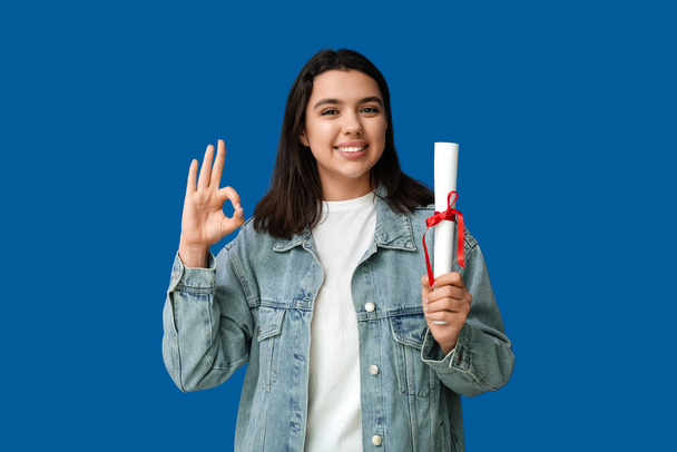 Счастливая улыбающаяся студентка с дипломом, показывающим хороший жест на синем фоне - Фото, изображение