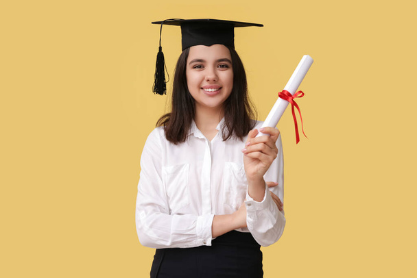Χαρούμενη χαμογελαστή φοιτήτρια με καπέλο αποφοίτησης με δίπλωμα σε κίτρινο φόντο - Φωτογραφία, εικόνα