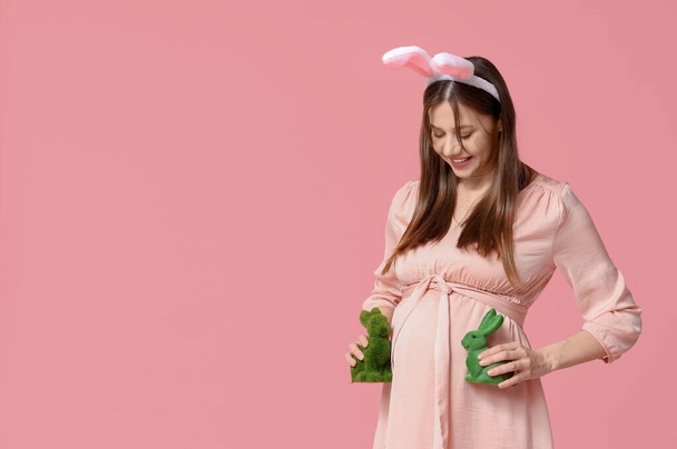 Beautiful young pregnant woman in bunny ears with rabbit toys on pink background. Easter celebration - Photo, Image