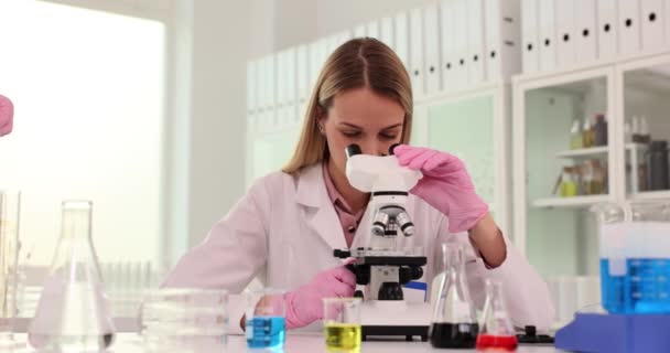 La científica mira en el microscopio mientras su colega trae estante con tubos de ensayo en el laboratorio. Mujeres tecnólogas estudian materiales alternativos y biotecnología - Metraje, vídeo