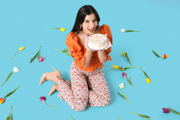 Young woman with sweet bento cake and beautiful tulips sitting on blue background. International Women's Day - Photo, Image
