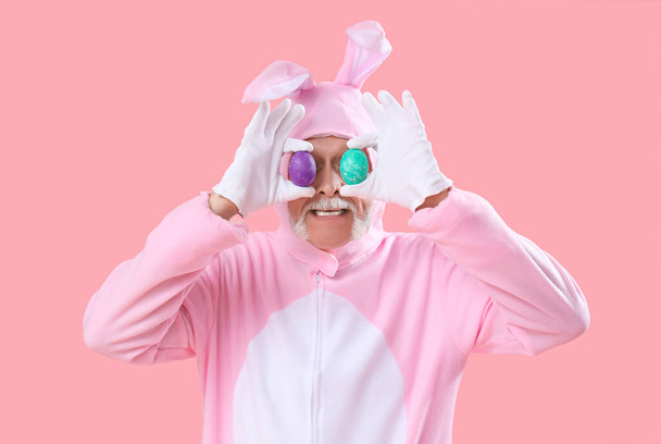 Senior mężczyzna w stroju królika pokrywa oczy pisankami na różowym tle - Zdjęcie, obraz