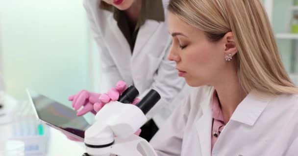 La mujer mira en el microscopio, mientras que el colega introduce datos en la hoja en la tableta en el laboratorio. Científicos calificados hacen pruebas con tecnología digital en la clínica - Imágenes, Vídeo