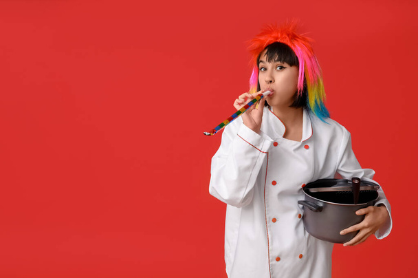 Funny young female chef with party blower and cooking pot on red background. Fool's day - Photo, Image
