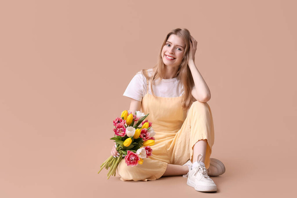Mooie jonge vrouw met boeket van mooie tulpen zittend tegen bruine achtergrond - Foto, afbeelding