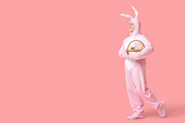 Счастливый молодой человек в костюме пасхального кролика держит корзину на розовом фоне - Фото, изображение