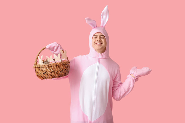 Feliz joven en traje de conejo de Pascua sosteniendo cesta y apuntando a algo sobre fondo rosa - Foto, imagen