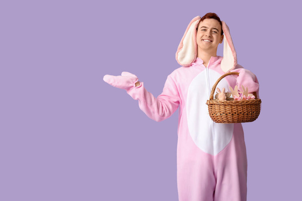 Счастливый молодой человек в костюме пасхального кролика держит корзину и указывает на что-то на сиреневом фоне - Фото, изображение