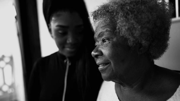 Pensive African American Senior žena zírá na dálku s kontemplativním pohledem, vnučka v pozadí ukazuje podporu pro babičku ve věku staré 80. let, černé a bílé - Fotografie, Obrázek