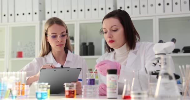 Vrouwelijke assistente toont lakmoesproef aan wetenschapper met papieren die hoofd knikken in het laboratorium. Vrouwelijke chemici stellen rapport op over experimenten met vloeistoffen - Video