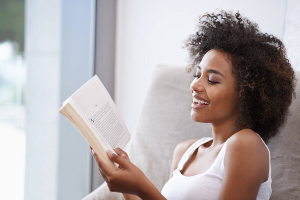 Mujer feliz, negra y leyendo con libro en sofá para novela, cuento o literatura en la sala de estar en casa. Persona femenina africana con sonrisa para información, noticias o conocimientos interesantes en la casa. - Foto, imagen