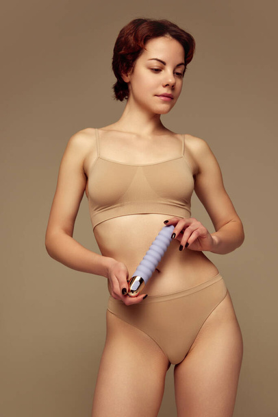 Tender young woman in cotton underwear holding vibrator against beige studio background. Intimacy and relaxation. Concept of female health care, toys for adults, sexuality, passion and pleasure - Photo, image
