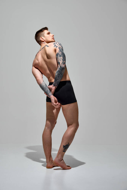 Zadní pohled portrét mladého tetovaného muže ve spodním prádle držícího ruce za zády na pozadí šedého studia. Rozumím. Koncept přírodní krásy, módy a stylu, mužského zdraví a péče o tělo. Reklama - Fotografie, Obrázek