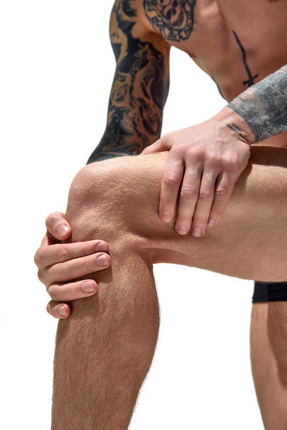 Κοντινό πλάνο του άνδρα με τατουάζ κρατά το χέρι στο γόνατο σε λευκό φόντο στούντιο. Πόνος στις αρθρώσεις. Αντιγραφή χώρου. Έννοια της φυσικής ομορφιάς, της μόδας και του στυλ, της ανδρικής υγείας και της φροντίδας του σώματος. ΠΑΡΑΡΤΗΜΑ II - Φωτογραφία, εικόνα
