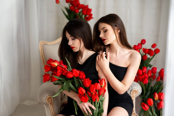 Красивая, милая, очаровательная, успешная пара девушек в платьях с макияжем, с красными тюльпанами в руках, обнимающимися, празднующими 8 марта, женский день, девичник или день рождения. Концепция ЛГБТ. - Фото, изображение
