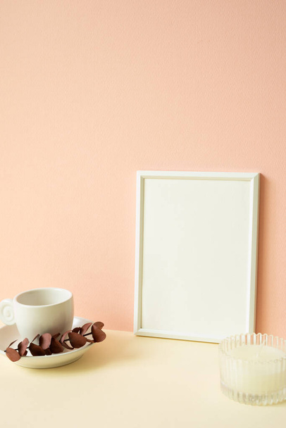 Біла порожня рамка картини з чашкою кави, свічка на стіл зі слонової кістки. рожевий фон стіни - Фото, зображення