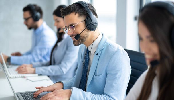 Operador de centro de llamadas guapo seguro que ayuda a los clientes, participa en la conversación, rodeado de colegas en la oficina del centro de llamadas. - Foto, imagen