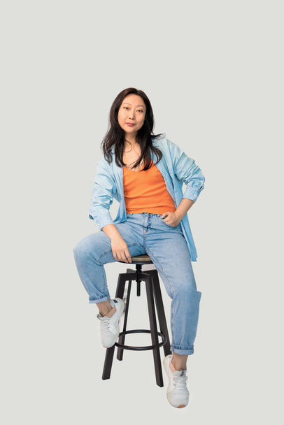Photo portrait full body view of Asian woman sitting on bar stool isolated on grey background - Foto, Imagen