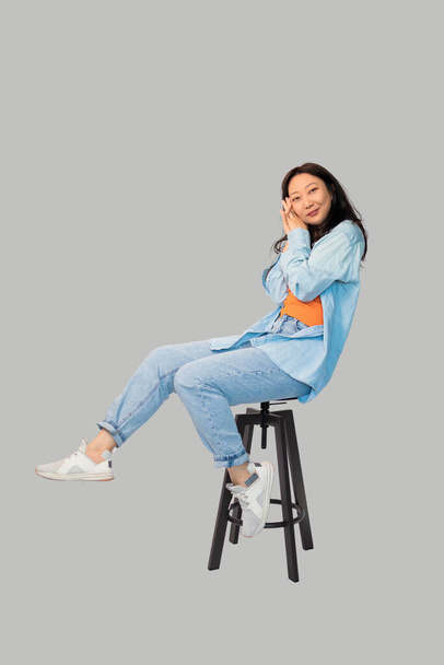 Ganzkörperansicht der attraktiven jungen Frau, die auf einem hohen Schemel sitzt. Vereinzelt auf grauem Hintergrund. - Foto, Bild